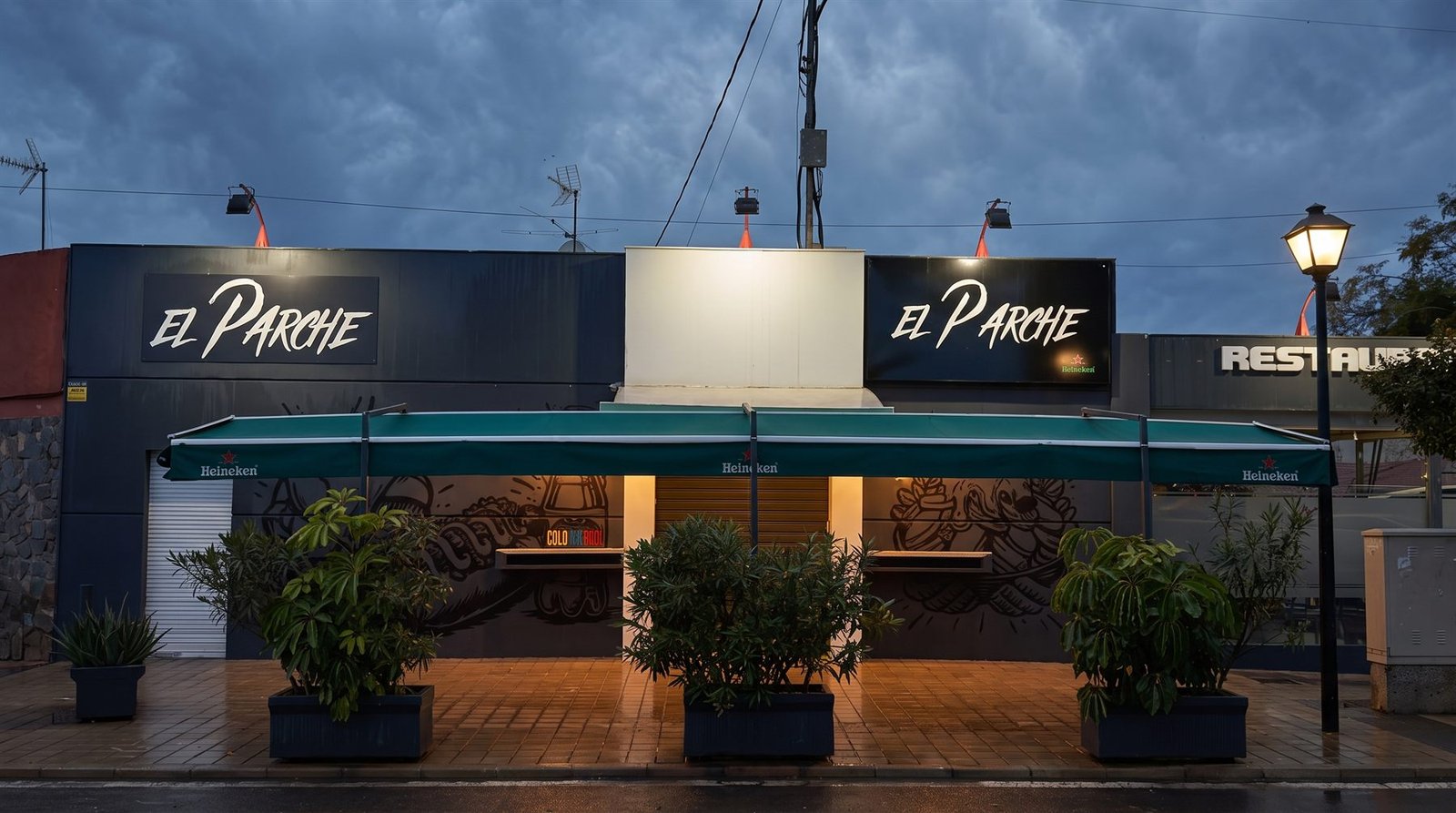 Storefront of El Parche Street Food in Alicante at sunset, with the awnings and the mural