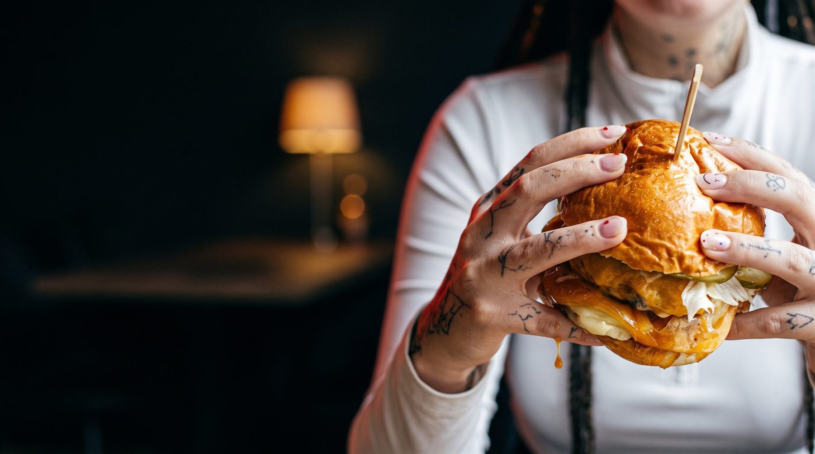 Two tattooed hands holding a burger with melted cheese dripping down